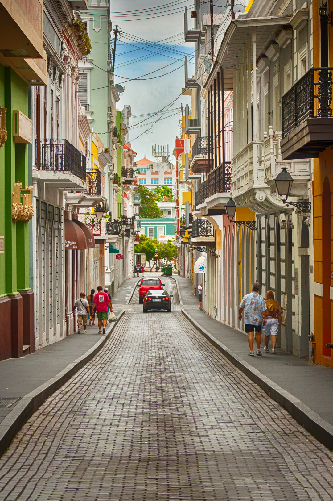 Brick almost empty street with many colorful buildings showing their balconies with Spanish colonial architecture influences.  a few people walking on the sidewalks and two cars on the background