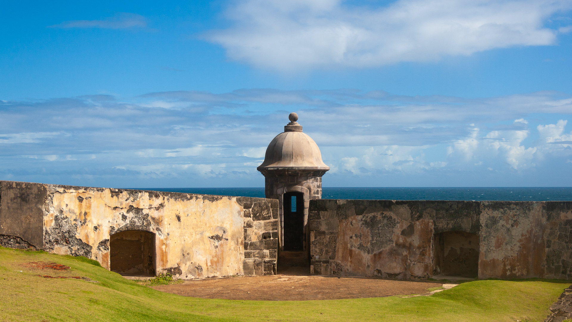 section of castle showing a sentry box with the ocean and blue skies with some clouds on background, green grass on the foreground.