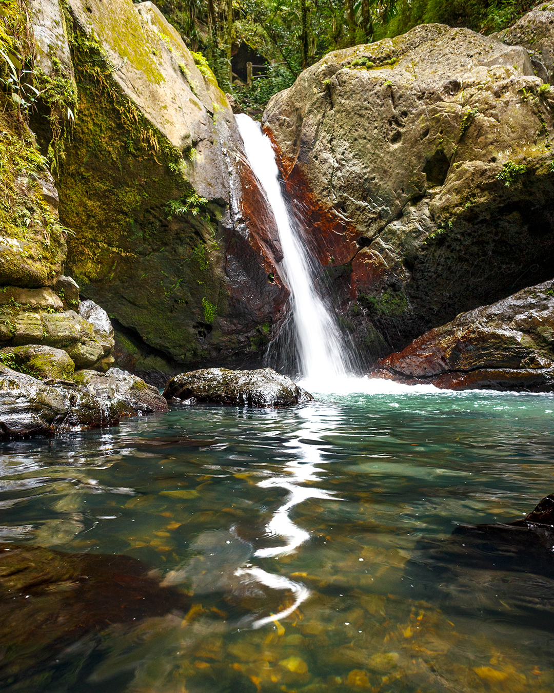 beautiful waterfall between two huge boulders with a reflection on the water of the waterfall, green tropical vegetation on the rocks and on the background.  river rocks on the bottom of the pond can be seen.