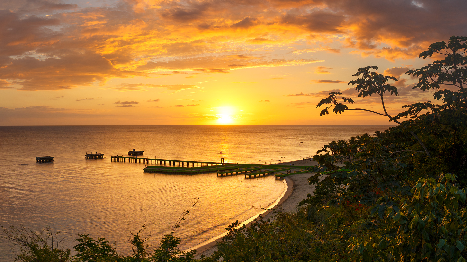 Landscape sunset scene of shoreline with dock, sun looks formless due to Saharan dust. Lovely orange and yellow hues, warm colors. Tropical lush on the foreground. 