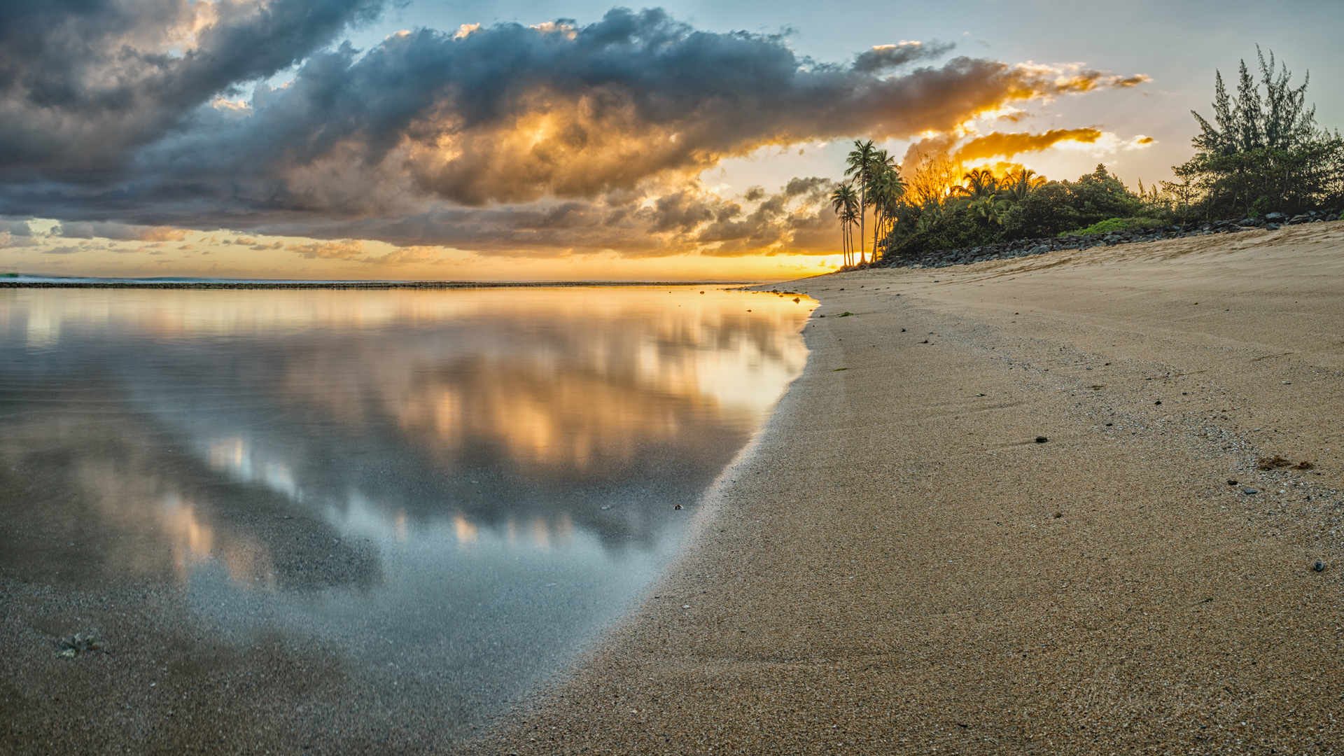 landscape of a golden sand beach with a very calm pond, a reflection of the clouds and skies can be seen in the water with beautiful orange and blue hues due to the sunrise. 