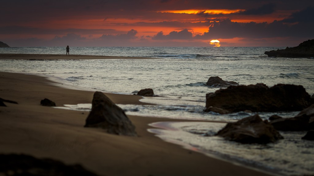 sunset scene, one person is standing on the sand contemplating the orange skies from the sunset, the scene is dark and there are rocks in the shore.