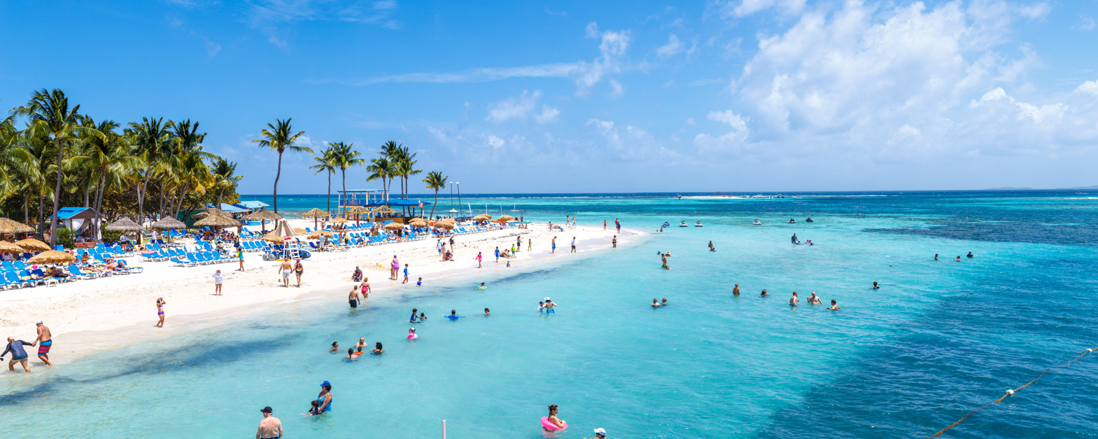 panoramic image of resort style beach with white sands, palms, clear blue waters with people having fun in the water and on the sand.  It's a sunny day and the skies are blue, there are blue beach chairs on the sand and bugaloos. 