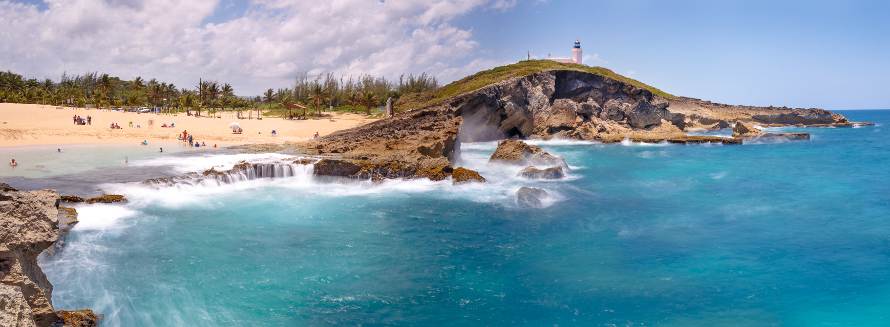 landscape of coastline with volcanic rocks, blue turquoise waters, waterfall like water movement, background include hill with lighthouse on top and people on the sand and beach.