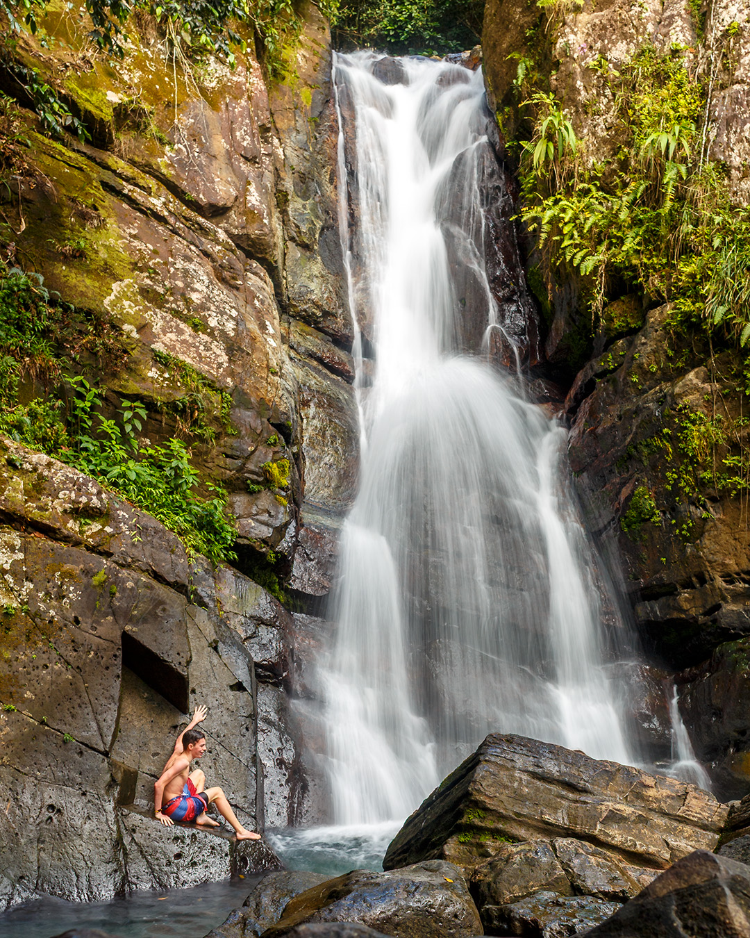 seated kid on a rock enjoying a very high waterfall with tropical green vegetation on rocks 